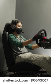 Handsome Asian Young Man Gamer Wearing Mask In Headphones With Pc Computer Playing Car Racing Video Game At Home And Steering Wheel