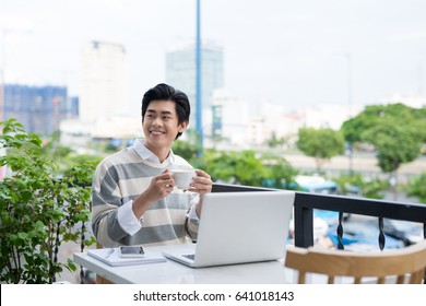 Handsome Asian Young Man Working On Laptop And Smiling While Enjoying Coffee In Cafe