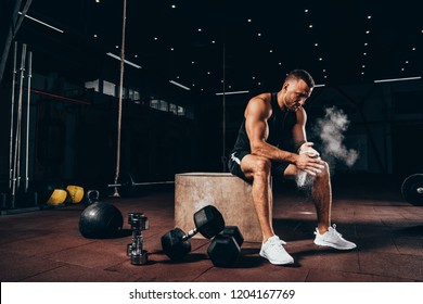 Handsome Athletic Man Sitting On Cube With Gym Equipment Around And Clapping Hands With Talc Before Workout