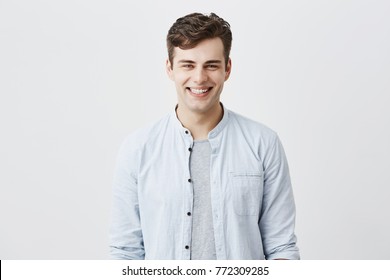 Handsome attractive young man dressed in light blue shirt over t-shirt with dark hair and appealing blue eyes looknig at camera, smiling broadly, demonstrating white teeth, being happy and pleased.
