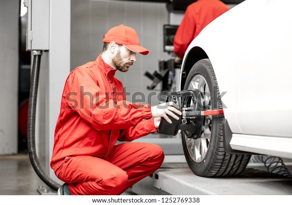 Handsome auto mechanic in red uniform fixing\
disk for wheel alignment at the car\
service