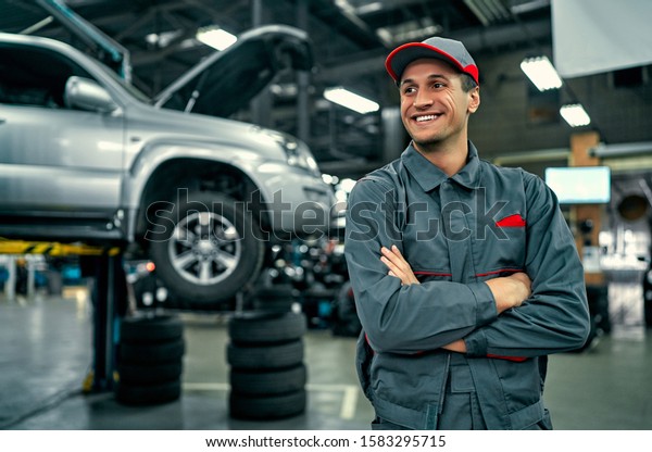 Handsome auto service mechanic in uniform is\
standing on the background of car with open hood, smiling and\
looking away. Car repair and\
maintenance.
