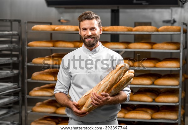 Handsome baker in uniform\
holding baguettes with bread shelves on the background at the\
manufacturing