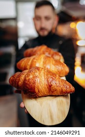 Handsome baker in uniform holding freshly baked croissants at the bakery - Shutterstock ID 2146049291