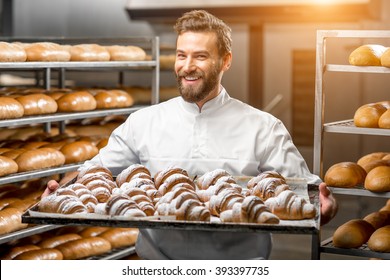 Handsome baker in uniform holding tray full of freshly baked croissants at the manufacturing - Shutterstock ID 393397735