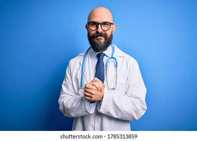 Handsome bald doctor man with beard wearing glasses and stethoscope over blue background with hands together and crossed fingers smiling relaxed and cheerful. Success and optimistic