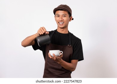 Handsome barista asian man wearing brown apron and black t-shirt isolated over white background . Barista holding milk jug and coffe cup practicing making coffee latte - Shutterstock ID 2227547823