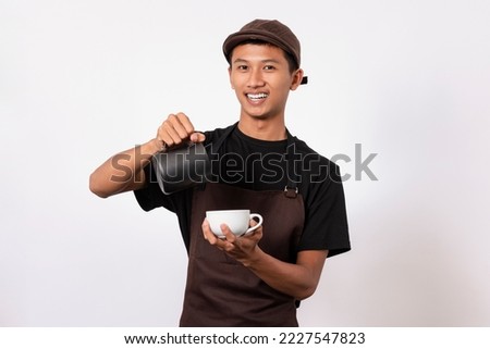 Handsome barista asian man wearing brown apron and black t-shirt isolated over white background . Barista holding milk jug and coffe cup practicing making coffee latte