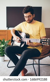 Handsome Beard Man Is Taking Online Guitar Course. Guy In A Yellow T-shirt Is Playing Acoustic Guitar At Home With Beautiful Interior. Doing Classes Of Chords While Staying On Quarantine