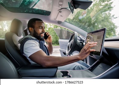 Handsome Bearded African Man Touches The Touchscreen In His New High-tech Electric Vehicle While While Talking By Phone And Smiling. Self Driving Vehicle Concept