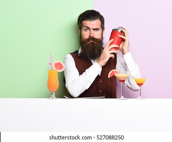 Handsome Bearded Barman With Long Beard And Mustache Has Stylish Hair On Serious Face Holding Shaker And Made Alcoholic Cocktail In Vintage Suede Leather Waistcoat On Purple Green Studio Background