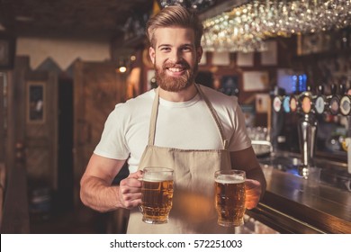 Handsome bearded bartender in apron is holding beer, looking at camera and smiling while standing near the bar counter in pub - Powered by Shutterstock