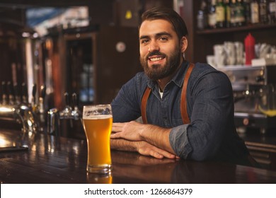 Handsome bearded bartender smiling joyfully to the camera, leaning on the bar counter, serving delicious beer at his pub, copy space. Brewer offering tasty craft beer at his restaurant - Powered by Shutterstock