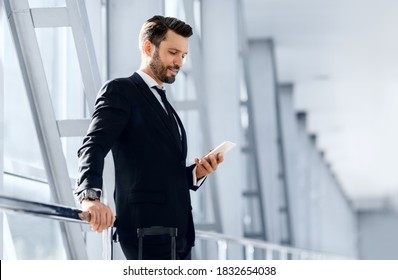 Handsome bearded businessman checking email on mobile phone while waiting for flight, standing next to window at airport, copy space. Middle-aged entrepreneur in stylish suit using smartphone