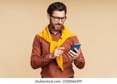 Handsome bearded businessman in formal shirt holding mobile phone reading text message, communication isolated on beige background. Smiling guy using mobile app, shopping online, ordering food 