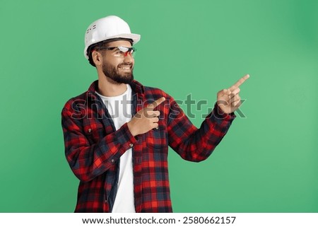 Handsome bearded engineer or constructor man in casual outfit pointing finger away over green background