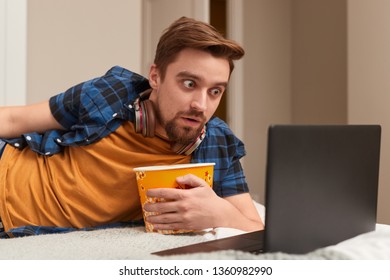 Handsome Bearded Guy With Bucket Of Popcorn Watching Interesting Film On Laptop With Astonishment While Lying On Bed During Movie Night At Home