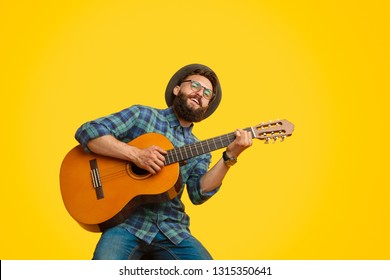 Handsome bearded guy in stylish hat and glasses smiling and playing acoustic guitar while sitting on bright yellow background - Powered by Shutterstock