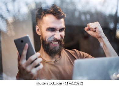 Handsome bearded hipster man sitting with laptop working online and having selling on photobank, freelance concept, portrait, online job, happy, victory sign, the winner. Sale, phone, technology