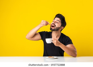 Handsome bearded Indian man eating healthy whole grain biscuits dipped in coffee of Tea or chai in a cup, sitting at table against yellow background