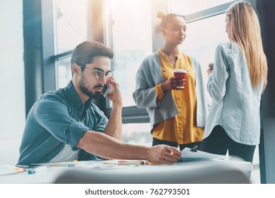 Handsome bearded male designer discusses new graphic project with colleague over smart phone, share opinions, sit in spacious office builduing. Two females chat about something with takeaway coffee - Shutterstock ID 762793501