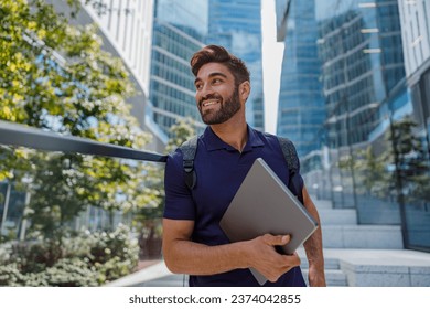 Handsome bearded male manager standing with laptop on skyscrapers background and looks away - Powered by Shutterstock