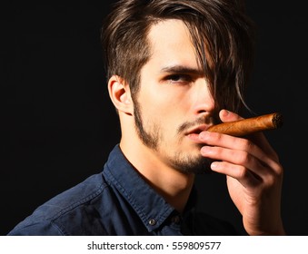 handsome bearded man in blue shirt with stylish hair on serious face smoking cigar in black studio background