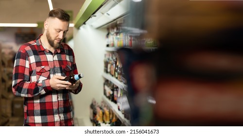 Handsome Bearded Man Chooses Beer In The Supermarket. Customer Buying Alcohol In A Liquor Store. Man Looking At Bottle Of Beer, Alcohol Purchase Concept, Banner, Place For Text