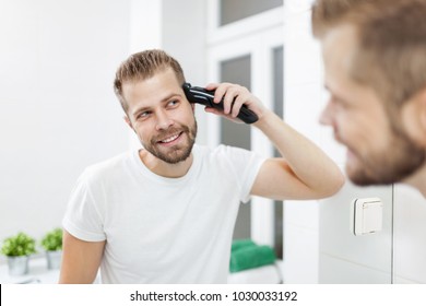 Handsome bearded man cutting his own hair with a clipper