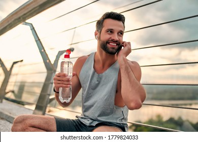 Handsome bearded man doing sport on the street. Having rest, drinking water and listening to music with wireless earphones after morning run - Powered by Shutterstock