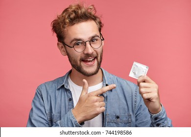 Handsome bearded man dressed casually, indicates at rubber condom, advertises new aromatic and qualified kind, recommends to buy it, guarantees good protection. Advertising concept. Contraception