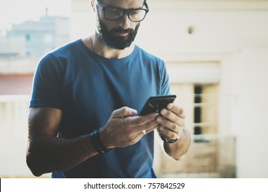 Handsome Bearded Man Holding Smartphone Hands And Touching Screen Of Modern Mobile Phone. Blurred Background.Blogger Using Mobile Gadget On The Balcony. Horizontal