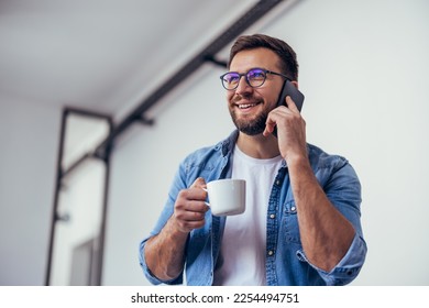 Handsome bearded man leaning on office desk, drinking coffee and talking on the phone. - Powered by Shutterstock