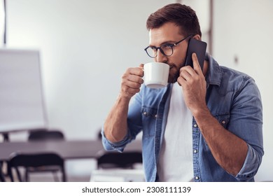 Handsome bearded man leaning on office desk, drinking coffee and talking on the phone. - Powered by Shutterstock