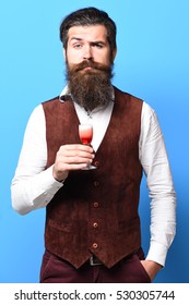 Handsome Bearded Man With Long Beard And Mustache On Serious Face Tasting Glass Of Alcoholic Shot In Vintage Suede Leather Waistcoat On Blue Studio Background