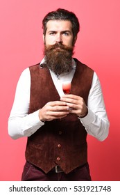 Handsome Bearded Man With Long Beard And Mustache On Serious Face Holding Glass Of Alcoholic Shot In Vintage Suede Leather Waistcoat On Red Studio Background