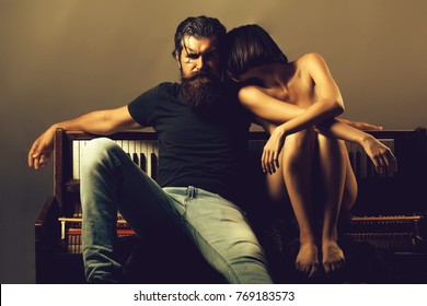 handsome bearded man with long beard on serious face near young woman or girl topless with sexy beautiful bare chest or breast on naked body and belly in panties near old wooden piano keyboard