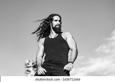 Handsome Bearded Man With Long Dark Hair. Fashionable Man With Long Wavy Hair And Strong Healthy Body. Mens Style And Wellness Concept. Strong Muscular Handsome Man At Blue Summer Sky Background