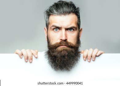 красивый бородатый человек с длинной пышной бородой и усами на удивленное лицо с белым листом бумаги в студии на сером фоне, копировать пространство