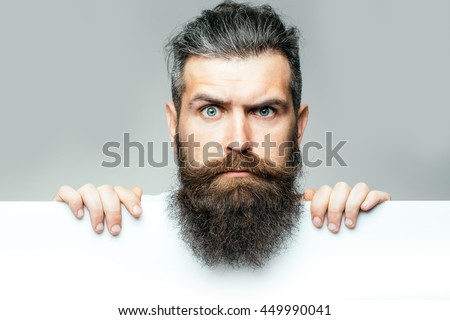 handsome bearded man with long lush beard and moustache on surprised face with white paper sheet in studio on grey background, copy space