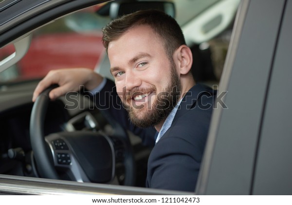 Handsome bearded man\
looking at camera from car cabin. Owner of automobile smiling,\
posing and holding hand on steering wheel. Concept of purchasing\
vehicles in car\
center.
