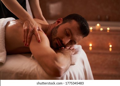 Handsome bearded man lying with closed eyes and an experienced massage therapist touching his shoulders