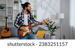 Handsome bearded man musician plays the guitar, takes pleasure of music and his hobby at ligt living room. Modern entertainment, music and happy young people concept.