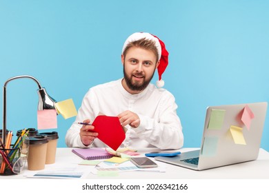 Handsome Bearded Man Office Worker In Santa Claus Hat Sitting At Workplace And Signing Postcards, Congratulating With Winter Holidays, Making Surprise. Indoor Studio Shot Isolated On Blue Background