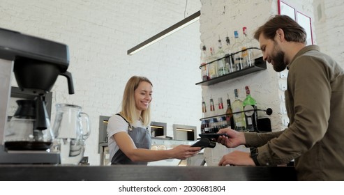 Handsome Bearded Man Paying For Takeaway Coffee With Mobile Phone At Coffee Shop. Worker Serving A Customer Who Uses Smartphone To Pay In Coffee Shop.