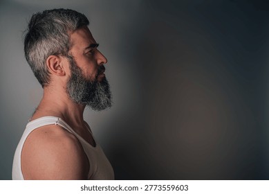 Handsome bearded man posing in studio multiple views A man stands in profile, looking to the side. He has a gray beard and short hair. He wears a simple white tank top and is in a well-lit indoor space.