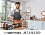 Handsome bearded man putting cut vegetables into frying pan in kitchen