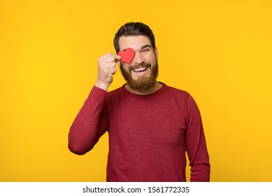 Handsome Bearded Man, In Red Sweater Fell In Love And Holding A Little Heart Over Eye, Like A Gesture, Standing Over Yellow Background 