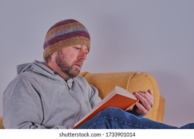 Handsome Bearded Man Sat In A Chair Reading A Book,there Is A Grey Background.