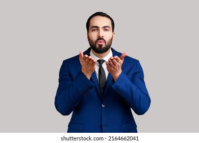 Handsome bearded man sending air kiss over palms, flirting to camera, demonstrating romantic amorous feelings, wearing official style suit. Indoor studio shot isolated on gray background.